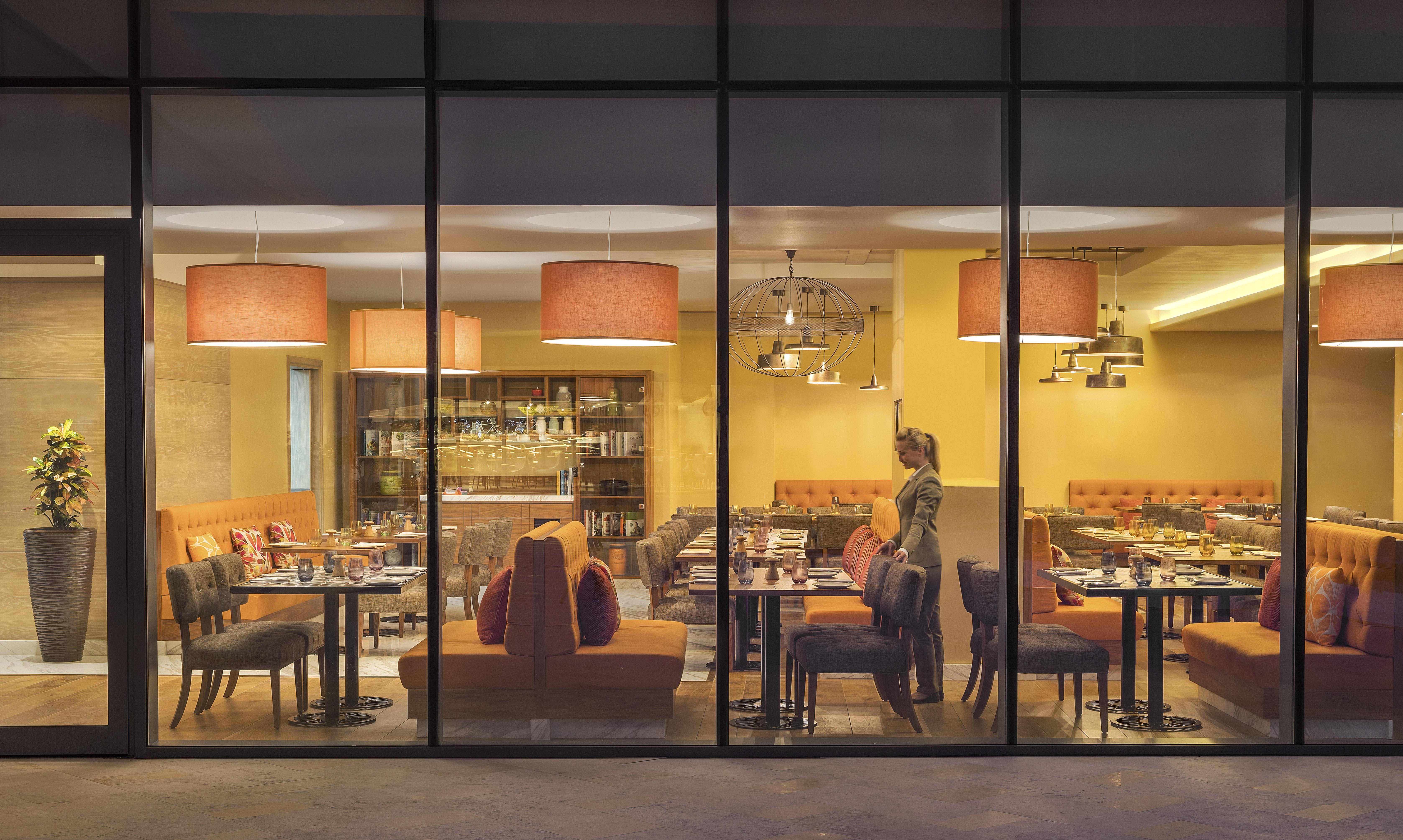 The Retreat Palm Dubai Mgallery By Sofitel Kültér fotó The photo shows a modern restaurant interior as viewed through large glass windows. Inside, there are several tables and chairs arranged, with some featuring orange upholstered seating. The decor includes stylish pendant lights hanging from the ceili