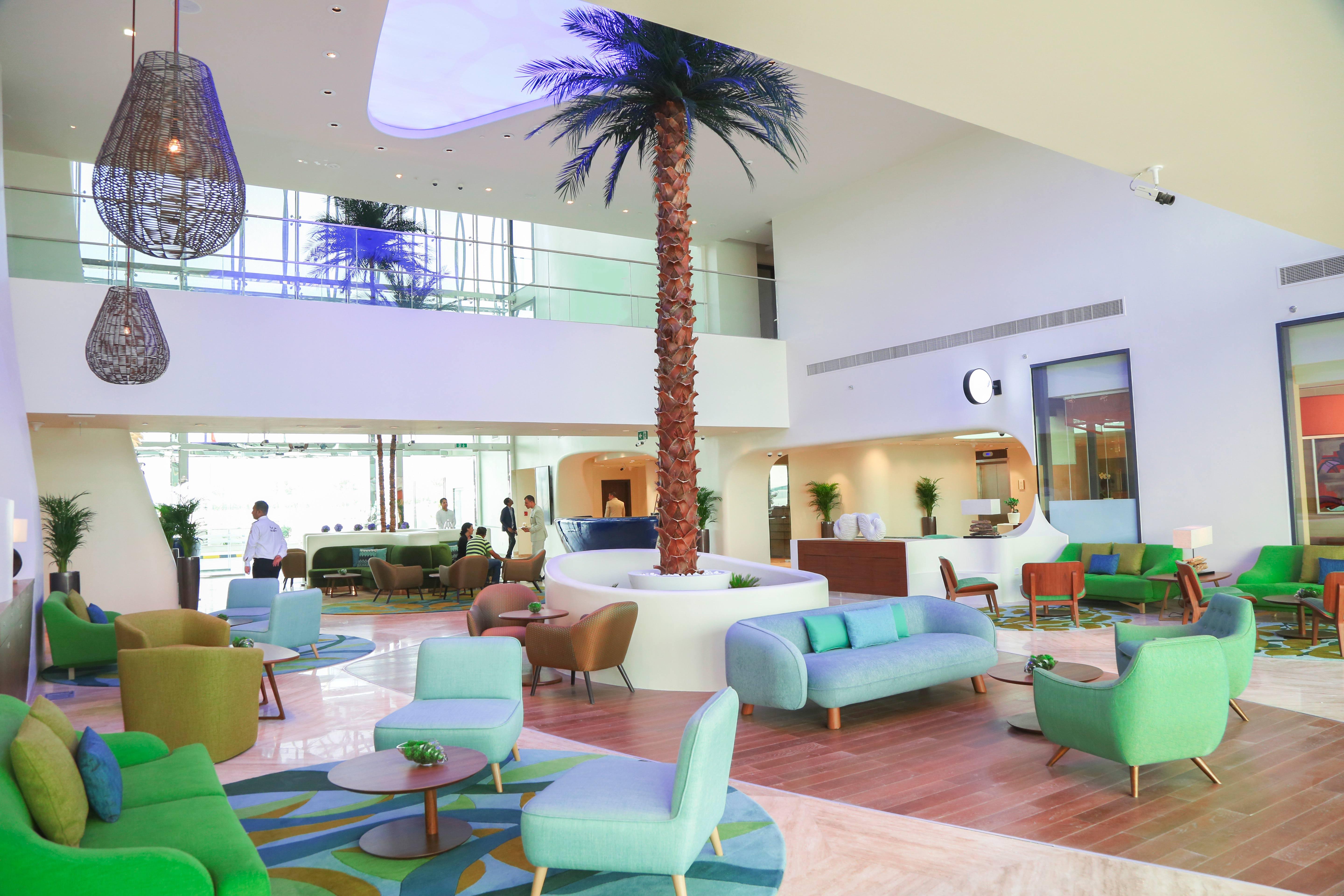 The Retreat Palm Dubai Mgallery By Sofitel Kültér fotó The photo shows a modern and stylish lobby area. It features a high ceiling with large windows allowing natural light to fill the space. In the center, there is a tall palm tree, adding a tropical feel. The seating consists of colorful couches and ch