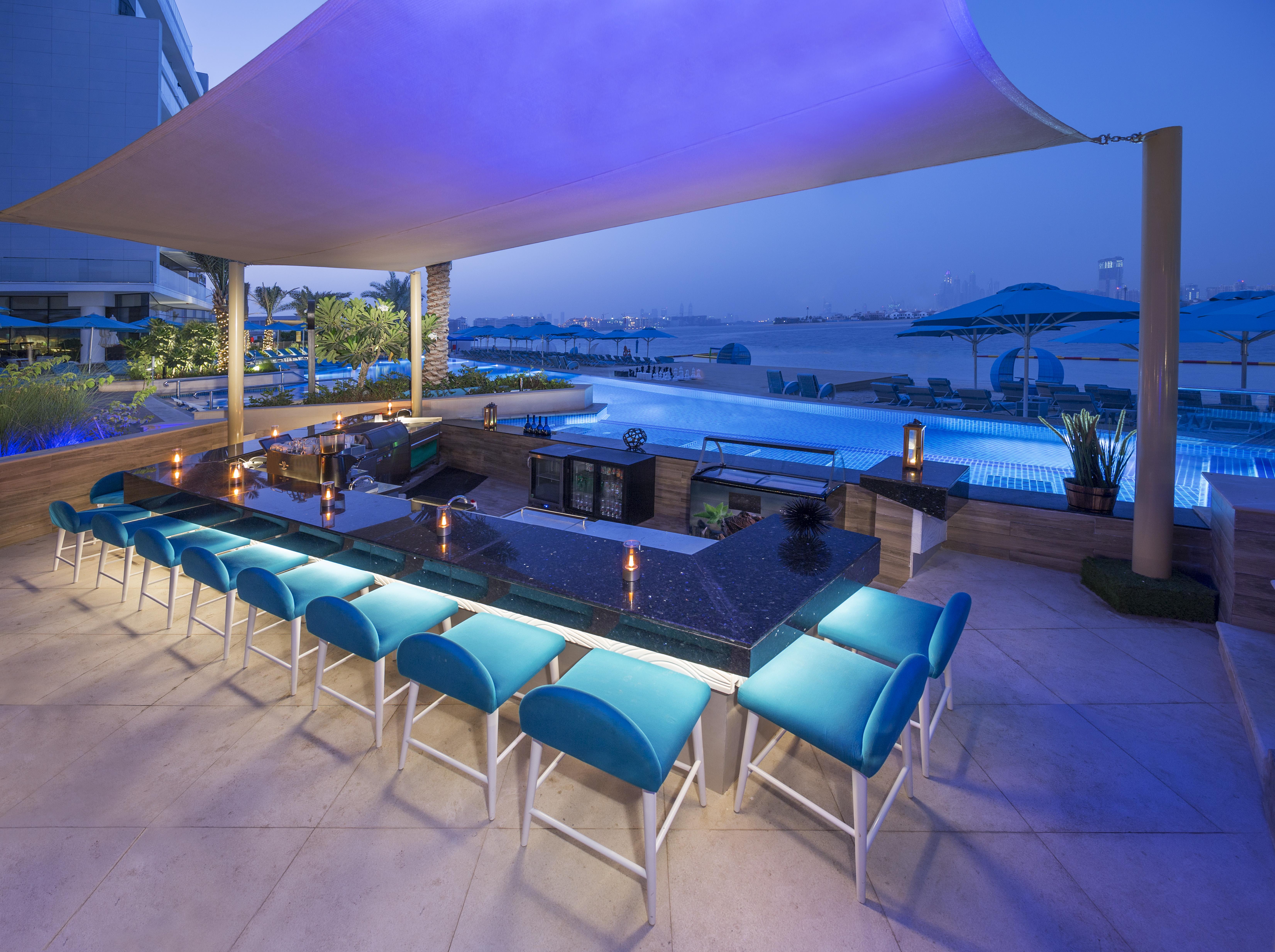 The Retreat Palm Dubai Mgallery By Sofitel Kültér fotó The photo depicts an elegant outdoor bar area beside a swimming pool. It features a long, dark countertop with several bar stools that have vibrant blue cushions. The setting is illuminated with soft lighting, creating a relaxed and inviting atmosphe