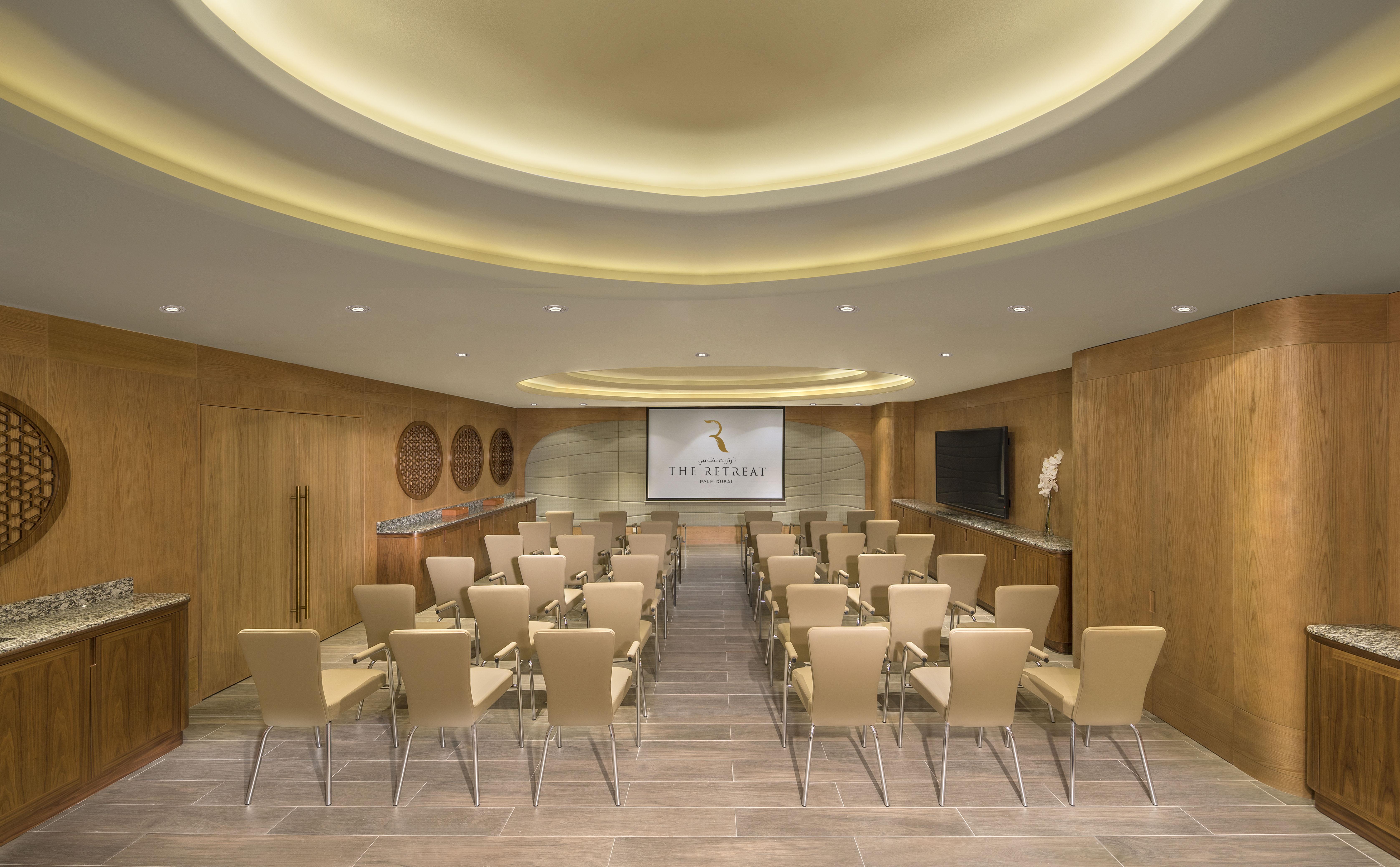 The Retreat Palm Dubai Mgallery By Sofitel Kültér fotó The photo shows a modern conference room. The room features a light wood and neutral decor with a curved ceiling that has soft lighting. There are rows of beige chairs arranged neatly facing a large screen on one wall. A sleek, wooden side area appea